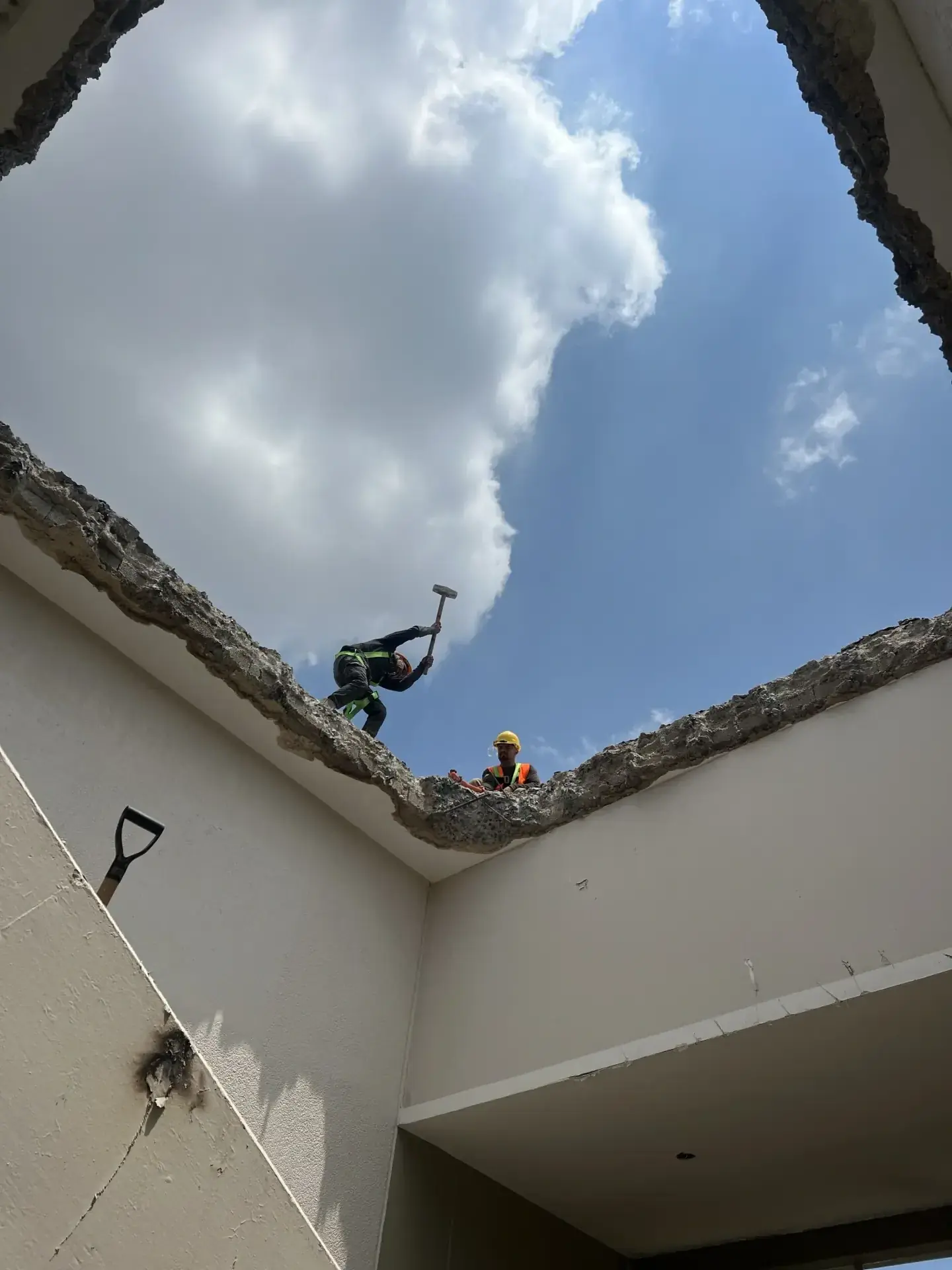 Dos obreros con casco y martillo demoliendo un techo de concreto, visto desde abajo, con cielo azul y nubes, en Guadalajara.